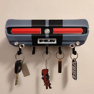 K&ouml;nnte beinhalten: Ein grau-schwarzer Schl&uuml;sselhalter im Shelby-Design mit roten R&uuml;ckleuchten und dem Wort "Shelby". Mehrere Schl&uuml;ssel und Schl&uuml;sselanh&auml;nger h&auml;ngen daran, darunter ein Deadpool-Schl&uuml;sselanh&auml;nger.