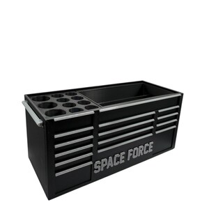 Op de afbeelding: Zwarte gereedschapskist met zilveren accenten en de woorden "SPACE FORCE" in zilver. De kist heeft meerdere laden en compartimenten voor gereedschapsopslag. Het bevat een sectie met ronde houders.