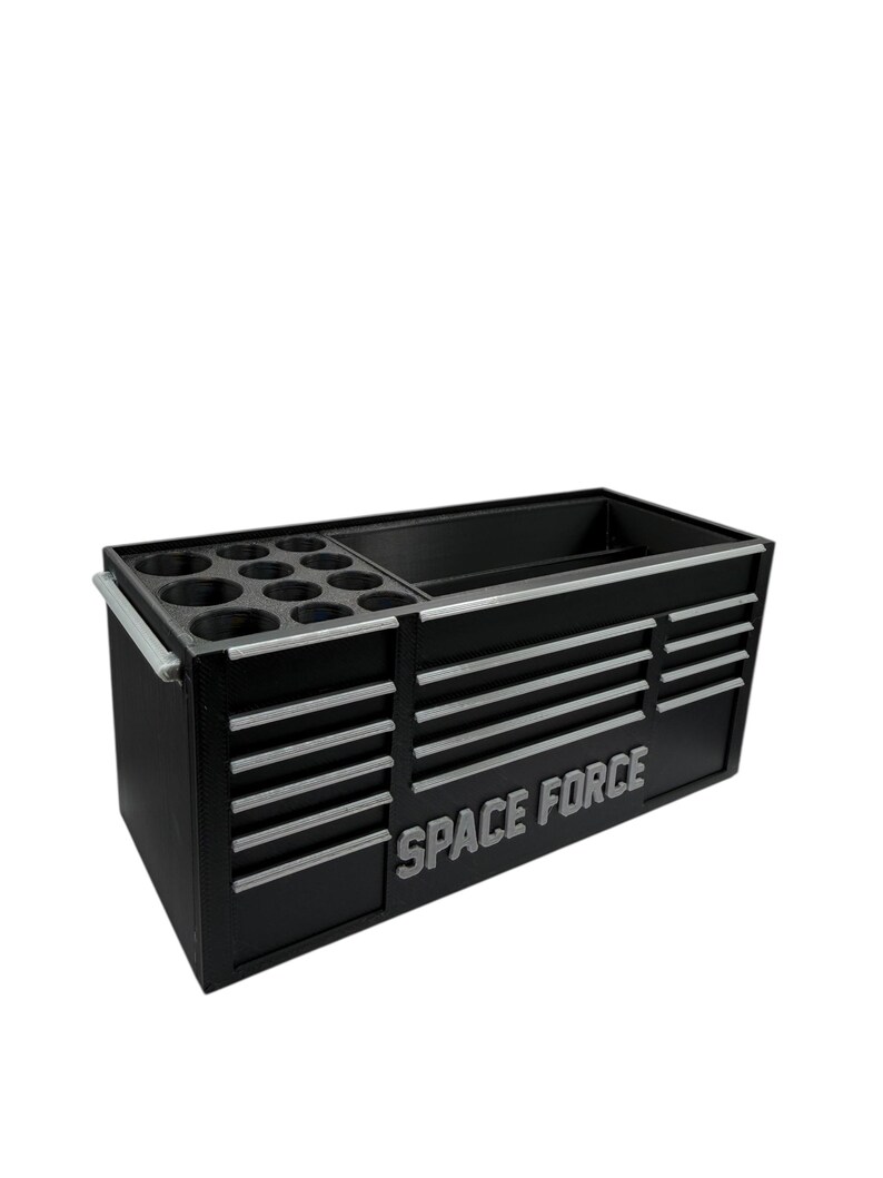 Op de afbeelding: Zwarte gereedschapskist met zilveren accenten en de woorden "SPACE FORCE" in zilver. De kist heeft meerdere laden en compartimenten voor gereedschapsopslag. Het bevat een sectie met ronde houders.