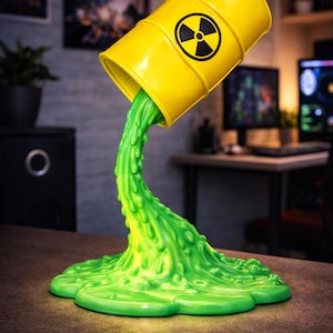 May include: A yellow barrel with a black radiation symbol is tilted, pouring out a stream of bright green, textured liquid. The liquid pools at the base, forming a unique decorative piece. Text reads: STANDS ON ITS OWN BY DESIGN.