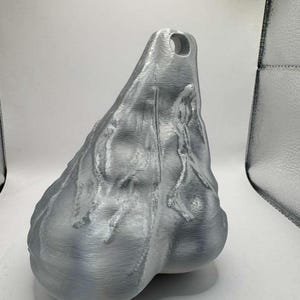 May include: A silver-colored, three-dimensional object with a textured, organic surface and a prominent, rounded shape at the base. The item features a tapered top section with a circular hole, displaying visible horizontal print lines throughout.