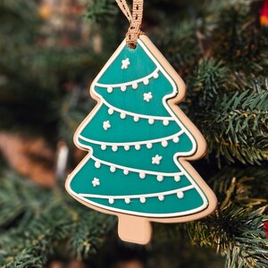 May include: A Christmas tree-shaped ornament with a teal green interior and white icing details. The ornament is hanging from a brown ribbon and is set against a backdrop of a Christmas tree.