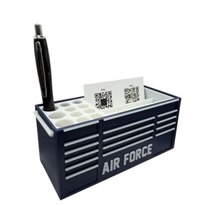 May include: A navy blue desk organizer shaped like a toolbox, with the words "AIR FORCE" in white. It has compartments for pens, business cards, and other small items. A black pen is in one of the holes.