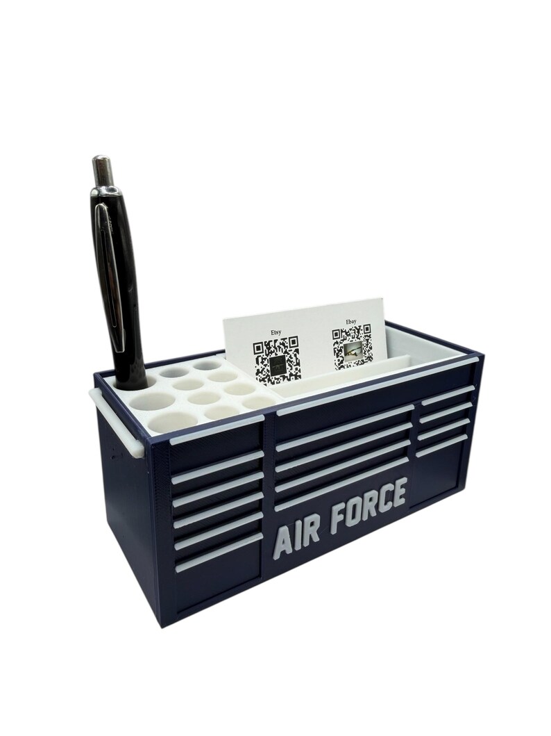 May include: A navy blue desk organizer shaped like a toolbox, with the words "AIR FORCE" in white. It has compartments for pens, business cards, and other small items. A black pen is in one of the holes.