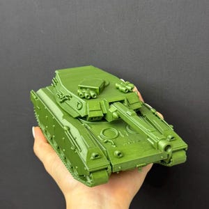 May include: A matte olive green plastic model tank featuring a rotating turret, a long main cannon, and detailed molded tracks. The military vehicle replica displays a textured surface finish with visible layered lines from the 3D printing process.