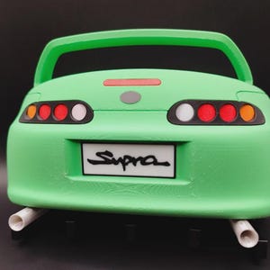 May include: A green model car with the word "Supra" on the license plate. The car has a spoiler, tail lights with red and white accents, and two exhaust pipes. The car is a toy or collectible.