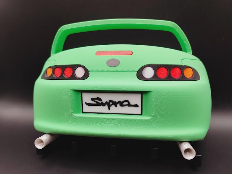 May include: A green model car with the word "Supra" on the license plate. The car has a spoiler, tail lights with red and white accents, and two exhaust pipes. The car is a toy or collectible.