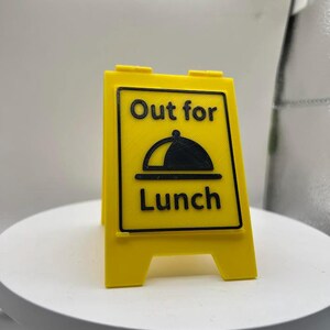 May include: A yellow, two-sided sign with the words "Out for Lunch" in black, along with a graphic of a covered dish. The sign is designed to stand upright, indicating a business is temporarily closed for lunch.