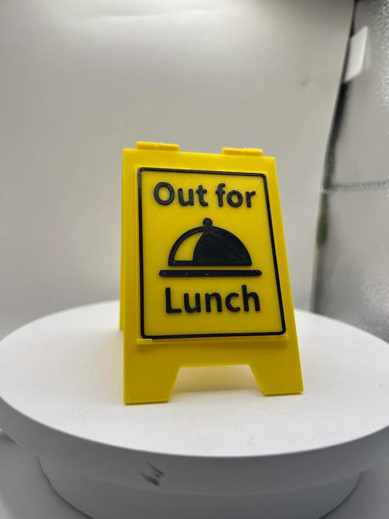 May include: A yellow, two-sided sign with the words "Out for Lunch" in black, along with a graphic of a covered dish. The sign is designed to stand upright, indicating a business is temporarily closed for lunch.