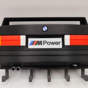 May include: Black wall-mounted key holder shaped like the rear of a classic BMW M3 car. Features red, white, and orange taillights, a BMW logo, and the "M Power" emblem. Includes four key hooks and dual exhaust pipes.