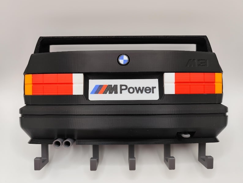 May include: Black wall-mounted key holder shaped like the rear of a classic BMW M3 car. Features red, white, and orange taillights, a BMW logo, and the "M Power" emblem. Includes four key hooks and dual exhaust pipes.