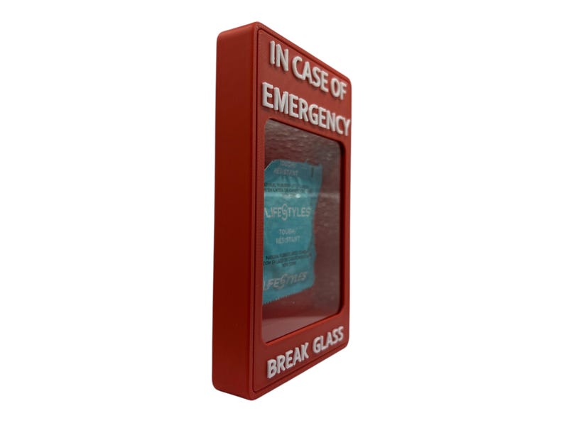 May include: A red rectangular box with the words "IN CASE OF EMERGENCY" and "BREAK GLASS" in white lettering. Inside the box is a blue package with the word "LIFESTYLES" visible.