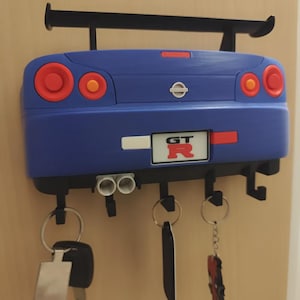 May include: A blue car-shaped key holder with a black spoiler and key hooks. The key holder has red and orange taillights and a "GT" license plate. Several keys hang from the hooks.