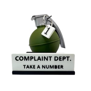 May include: A novelty desk sign with a green grenade-shaped object and the words "COMPLAINT DEPT. TAKE A NUMBER" in black text on a white background. A small tag with the number 1 hangs from the grenade.
