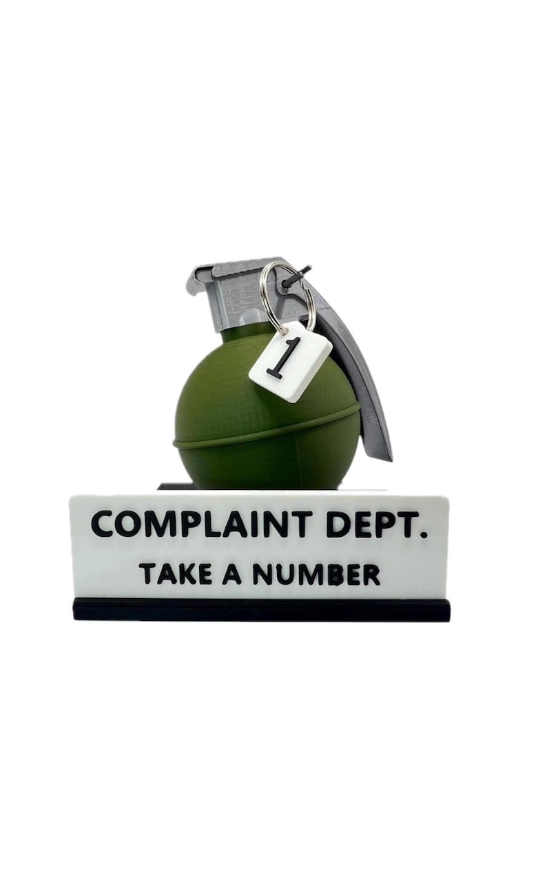 May include: A novelty desk sign with a green grenade-shaped object and the words "COMPLAINT DEPT. TAKE A NUMBER" in black text on a white background. A small tag with the number 1 hangs from the grenade.