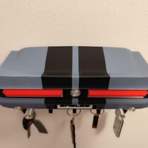 K&ouml;nnte beinhalten: Ein Wand-Schl&uuml;sselhalter in Form des Hecks eines klassischen Autos. Der Schl&uuml;sselhalter ist hellblau lackiert und hat schwarze Rennstreifen und rote R&uuml;cklichter. Das Wort "Shelby" ist auf dem Nummernschild zu sehen.