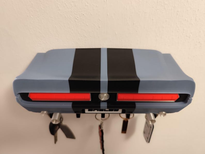 K&ouml;nnte beinhalten: Ein Wand-Schl&uuml;sselhalter in Form des Hecks eines klassischen Autos. Der Schl&uuml;sselhalter ist hellblau lackiert und hat schwarze Rennstreifen und rote R&uuml;cklichter. Das Wort "Shelby" ist auf dem Nummernschild zu sehen.