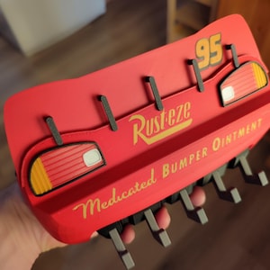 May include: A red wall-mounted hook rack shaped like Lightning McQueen's bumper from the movie Cars. The rack features the number 95, the 'Rusteze' logo, and the text 'Medicated Bumper Ointment'. It has several black hooks.