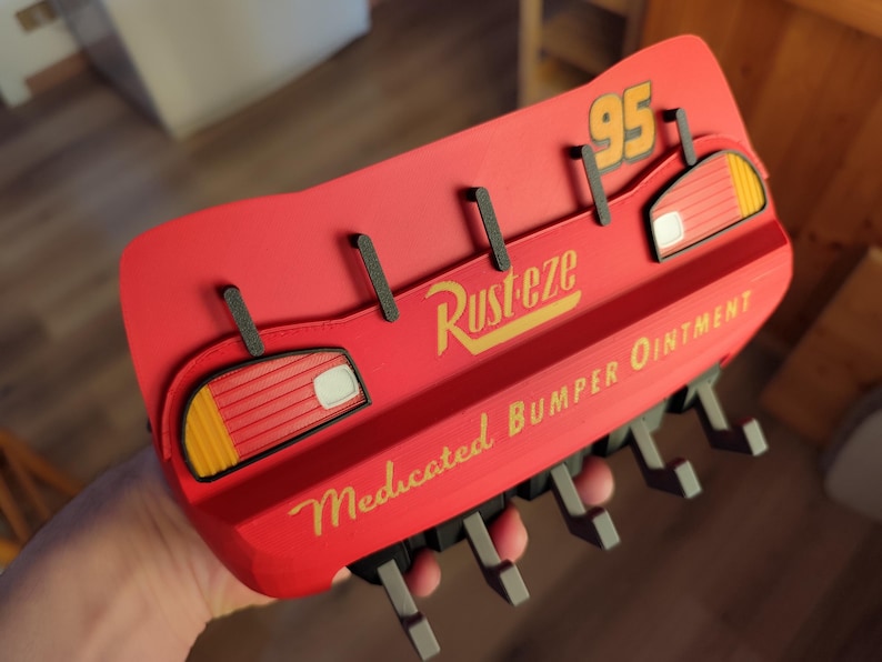 May include: A red wall-mounted hook rack shaped like Lightning McQueen's bumper from the movie Cars. The rack features the number 95, the 'Rusteze' logo, and the text 'Medicated Bumper Ointment'. It has several black hooks.