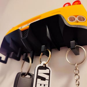 Puede incluir: Un llavero con forma de coche amarillo y negro montado en una pared blanca. El llavero tiene un dise&ntilde;o trasero de coche con dos tubos de escape y tres ganchos para llaves. Llaveros con llaves cuelgan de los ganchos.