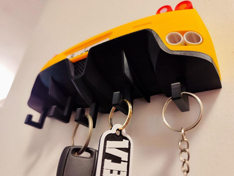 Puede incluir: Un llavero con forma de coche amarillo y negro montado en una pared blanca. El llavero tiene un dise&ntilde;o trasero de coche con dos tubos de escape y tres ganchos para llaves. Llaveros con llaves cuelgan de los ganchos.