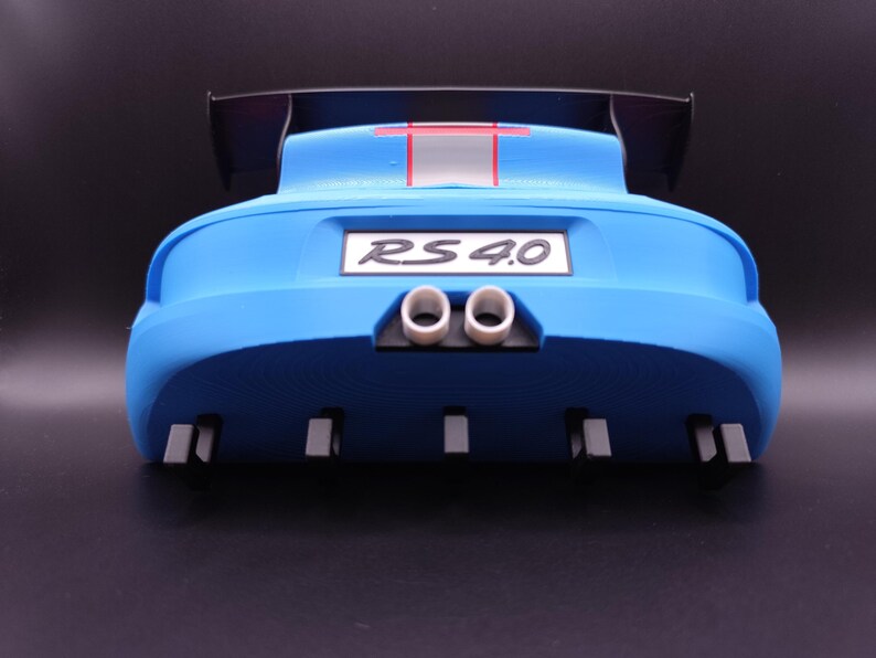 K&ouml;nnte beinhalten: Heck eines blauen Spielzeugautos mit einem schwarzen Spoiler. Das Auto hat ein wei&szlig;es Nummernschild mit der Aufschrift "RS 4.0". Es hat zwei wei&szlig;e Auspuffrohre und mehrere graue rechteckige Details. Ein roter Streifen akzentuiert die Oberseite.