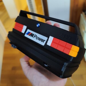 May include: Black model car rear end with red, white, and orange tail lights. The model has the BMW logo and the text "M Power". The model is held in a person's hand.