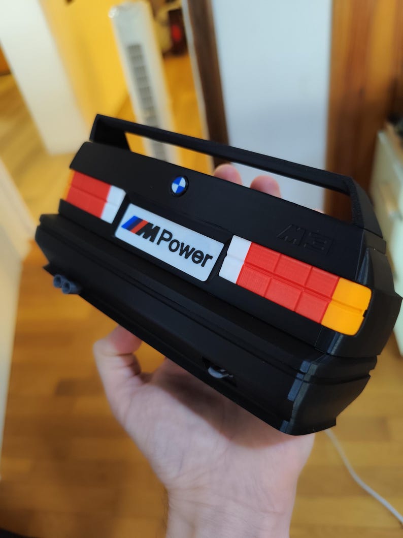 May include: Black model car rear end with red, white, and orange tail lights. The model has the BMW logo and the text "M Power". The model is held in a person's hand.