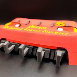 May include: A red 3D-printed Lightning McQueen bumper with the text "Rusteze Medicated Bumper Ointment" in yellow. The bumper has gray hooks and the number "95" in yellow.
