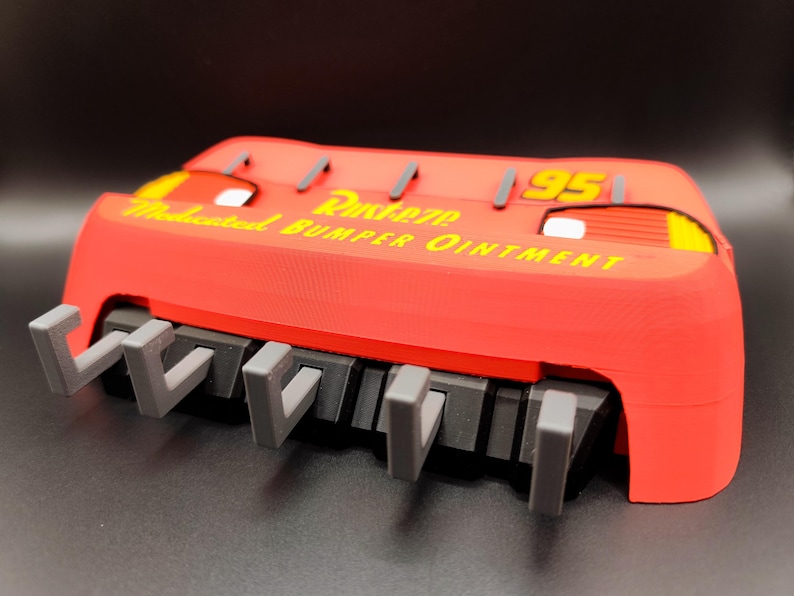 May include: A red 3D-printed Lightning McQueen bumper with the text "Rusteze Medicated Bumper Ointment" in yellow. The bumper has gray hooks and the number "95" in yellow.