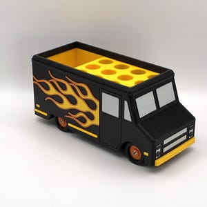 May include: A black food truck-shaped desk organizer with yellow flames on the side. The truck has a yellow interior with multiple circular compartments. The wheels and trim are orange and yellow.