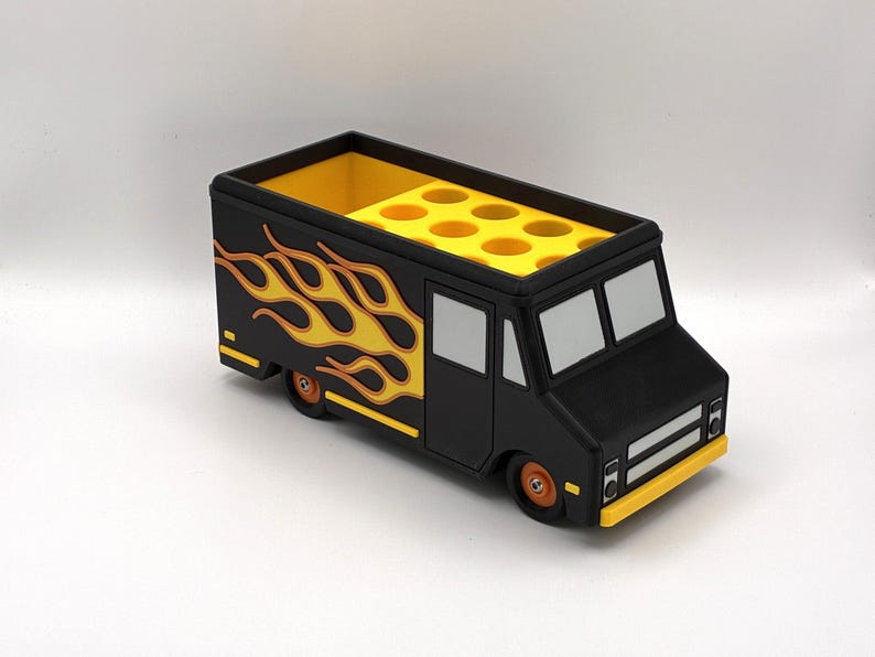 May include: A black food truck-shaped desk organizer with yellow flames on the side. The truck has a yellow interior with multiple circular compartments. The wheels and trim are orange and yellow.