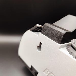 Pode incluir: Close-up de um dispositivo de pl&aacute;stico branco com uma superf&iacute;cie texturizada. O dispositivo tem um componente de espuma cinza escuro e um pequeno buraco de fechadura. A palavra "BECC" est&aacute; em relevo na lateral. O fundo &eacute; cinza escuro.
