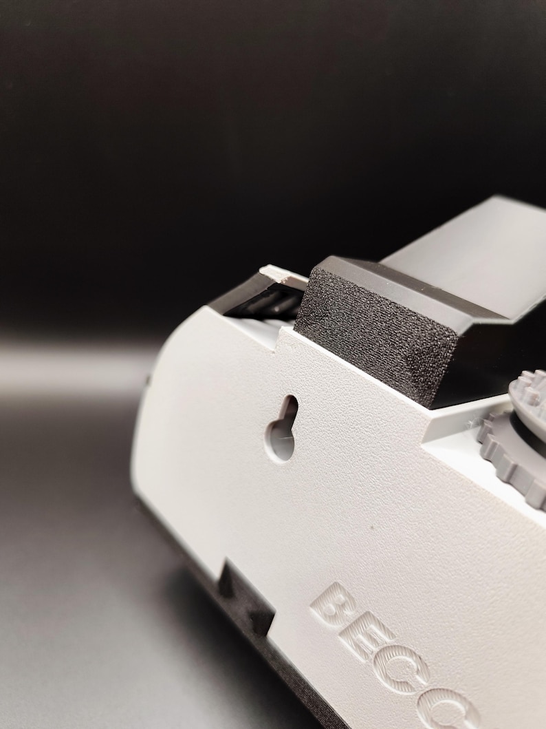 Pode incluir: Close-up de um dispositivo de pl&aacute;stico branco com uma superf&iacute;cie texturizada. O dispositivo tem um componente de espuma cinza escuro e um pequeno buraco de fechadura. A palavra "BECC" est&aacute; em relevo na lateral. O fundo &eacute; cinza escuro.