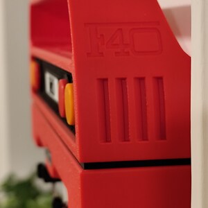 May include: A bright red model car component with the number "F40" embossed on the side. The component has a rectangular shape with vertical lines and orange and yellow accents. The object is positioned against a white background.