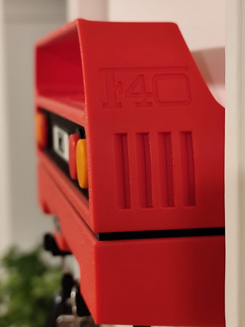 May include: A bright red model car component with the number "F40" embossed on the side. The component has a rectangular shape with vertical lines and orange and yellow accents. The object is positioned against a white background.
