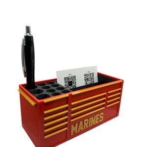 May include: Red and gold desk organizer with the word "MARINES" in gold lettering. The organizer has a pen holder, business card slot, and several small holes. A black pen and business cards are in the organizer.