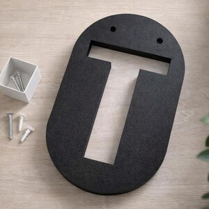 Op de afbeelding: Een zwarte, ovaalvormige letter "T" van textuurmateriaal, met twee kleine gaatjes aan de bovenkant. Een klein wit doosje met zilveren schroeven en drie extra schroeven staat naast de letter. Decoratief groen is op de achtergrond.