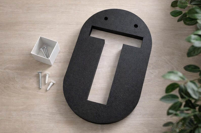 Op de afbeelding: Een zwarte, ovaalvormige letter "T" van textuurmateriaal, met twee kleine gaatjes aan de bovenkant. Een klein wit doosje met zilveren schroeven en drie extra schroeven staat naast de letter. Decoratief groen is op de achtergrond.