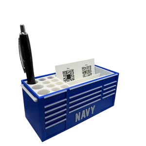 May include: A navy blue desk organizer shaped like a toolbox, with the word "NAVY" printed on the side. It has a section for pens, business cards, and a black pen. The organizer is designed to hold office supplies.