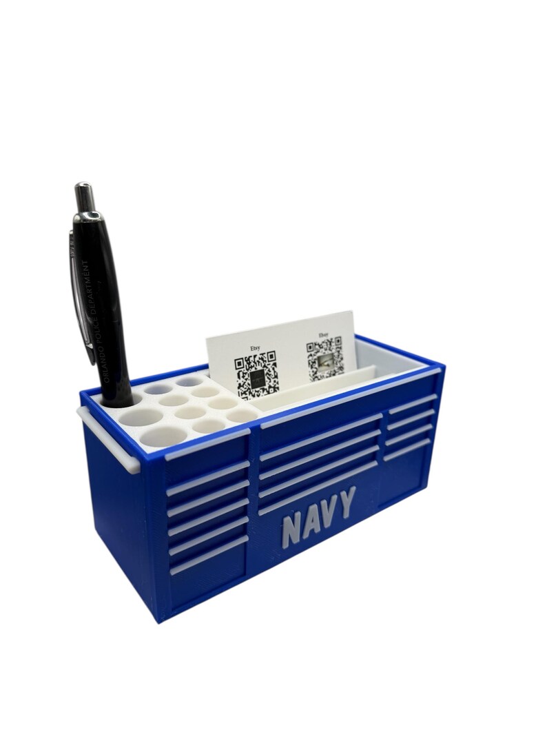 May include: A navy blue desk organizer shaped like a toolbox, with the word "NAVY" printed on the side. It has a section for pens, business cards, and a black pen. The organizer is designed to hold office supplies.