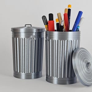 May include: Two silver metal trash cans with lids. One trash can is empty and the other is filled with various writing utensils, including pencils, pens, and markers.