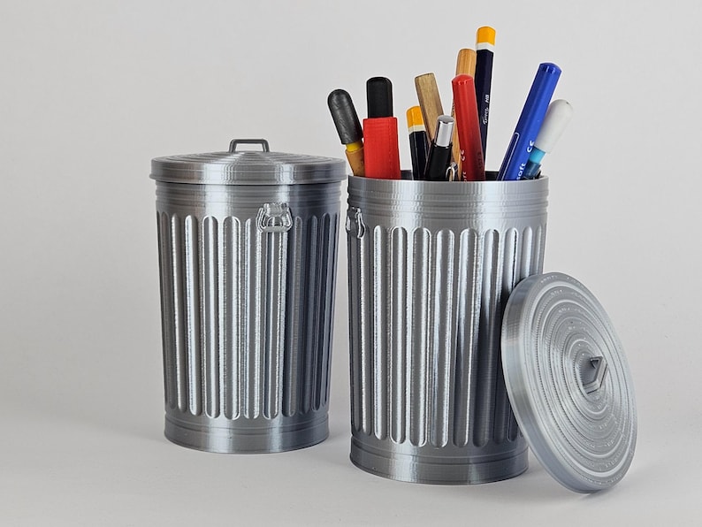 May include: Two silver metal trash cans with lids. One trash can is empty and the other is filled with various writing utensils, including pencils, pens, and markers.