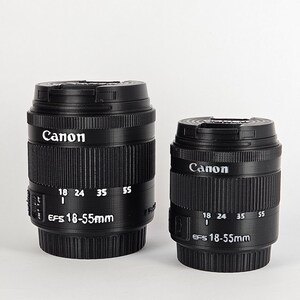 May include: Two black Canon EF-S 18-55mm camera lenses. The larger lens is in the background and the smaller lens is in the foreground.