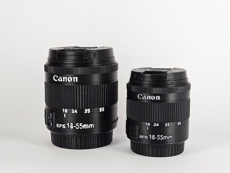 May include: Two black Canon EF-S 18-55mm camera lenses. The larger lens is in the background and the smaller lens is in the foreground.