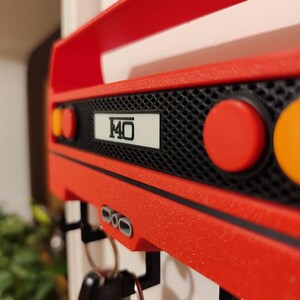 May include: A red key holder shaped like the rear of a sports car, with the number "F40" on a white plate. The key holder has black hooks for keys and round red and orange lights. The key holder is mounted on a white wall.