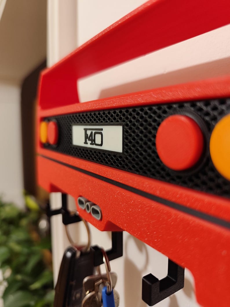 May include: A red key holder shaped like the rear of a sports car, with the number "F40" on a white plate. The key holder has black hooks for keys and round red and orange lights. The key holder is mounted on a white wall.