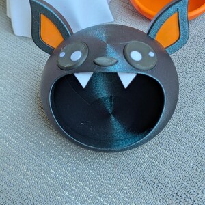May include: A dark gray bat-shaped container with orange-lined ears and white fangs. The container has large, round eyes with white highlights. The bat's mouth is open, forming a large opening. Other Halloween-themed containers are visible in the background.