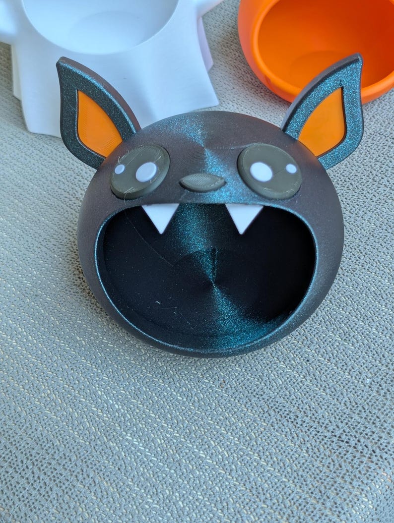 May include: A dark gray bat-shaped container with orange-lined ears and white fangs. The container has large, round eyes with white highlights. The bat's mouth is open, forming a large opening. Other Halloween-themed containers are visible in the background.