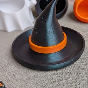 May include: A black witch hat-shaped trinket dish with an orange band, moon, and star cutouts. Other Halloween-themed containers are in the background, including a ghost, bat, and pumpkin.